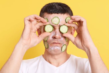 Beyaz kilden maskeli yakışıklı esmer adam gözlerini iki dilim salatalıkla kapatıyor. Erkekler için kaplıca, gençleştirme tedavileri, sarı arka planda izole edilmiş pozlar..