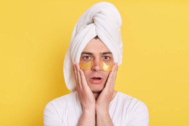 Şok olmuş bir adam kafaya havlu sarmış sabah kozmetik ameliyatları yapıyor, avuçlarını yanaklarında tutuyor, büyük gözlerle kameraya bakıyor, sarı arka planda izole edilmiş göz altı yamalarıyla poz veriyor..