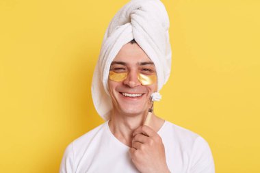 Yüzüne havlu sarılmış gülümseyen bir adam sabah kozmetik ameliyatları yapıyor, göz altında yamalarla poz veriyor, sarı arka planda izole edilmiş tekerlekli sandalyeyle yüz masajı yapıyor..