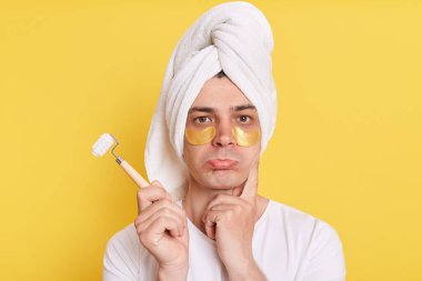Kafkasyalı üzgün bir adamın portresi. Kafasına havlu sarıp sabah kozmetoloji prosedürleri uyguluyor. Dudakları büzük vaziyette duruyor, sarı arka planda izole edilmiş göz altı yamalarıyla poz veriyor..