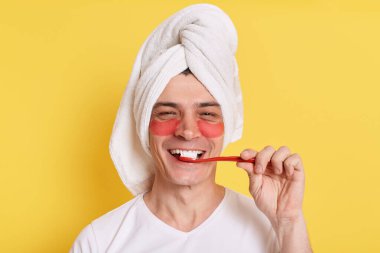Yüzüne havlu sarıp sabah kozmetoloji prosedürleri uygulayan, dişlerini fırçalayan, sarı arka planda izole edilmiş göz altı bantlarıyla poz veren olumlu gülümseyen beyaz adam..