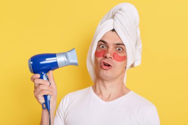 Kafkasyalı komik bir adam kafaya havlu sarmış sabah kozmetoloji prosedürleri uyguluyor, sarı arka planda izole edilmiş göz bantlarıyla poz veriyor, elinde saç kurutma makinesi ve şaşırmış bir ifade var..