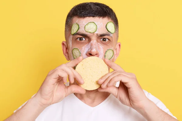 Indoor shot of serious man with dark hair applying mask with cucumber, holding sponge and covering mouth, looking at camera with strict expression, posing isolated over yellow background.