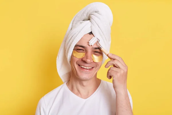 阳性男子用毛巾包裹在头上 做早间面部按摩 用眼底补丁按摩脸颊 进行美容术 用黄色背景隔开 — 图库照片