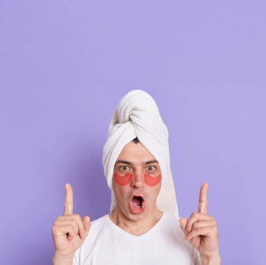 Şok olmuş bir adamın portresi mor arkaplan üzerinde izole edilmiş, büyük gözleri ve ağzı açık bir şekilde kameraya bakıyor, fotokopi alanını işaret ediyor, gözlerinin altındaki yamalarla poz veriyor..