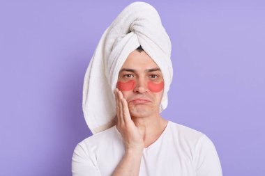 Havlu giymiş üzgün genç adamın portresi mor arka planda izole edilmiş göz kırışıklığı önleyici bantlar kullanıyor, üzüntüyü ifade ediyor, somurtkan dudaklarla kameraya bakıyor..