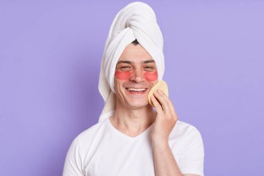 Havlu giymiş genç bir adamın portresi krem sürmek için göz kırışıklık önleyici bantlar ve sünger kullanır, kameraya olumlu bir ifadeyle bakar, sabah ameliyatlarından zevk alır..