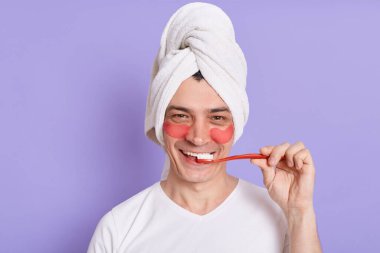 Ellerinde diş fırçası olan ve havluya sarılı bir adamın fotoğrafı, göz altı yamalarıyla dişlerini fırçalarken, sabah prosedürleri yaparken, mor arka planda izole bir şekilde poz verirken..