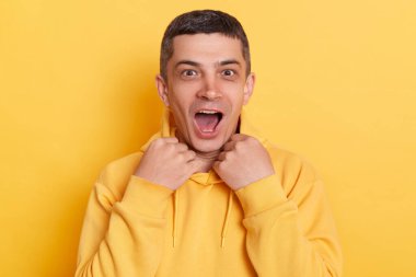 Şok olmuş yakışıklı bir adam sarı arka planda izole edilmiş bir şekilde poz veriyor, ağzı açık ve büyük gözlerle kameraya bakıyor, şaşkınlığını dile getiriyor..