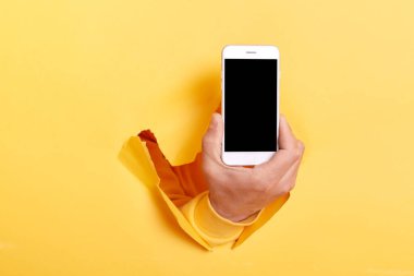 Mobile App, great offer. Male hand holding smartphone with white empty screen showing device close up to camera breaking through yellow paper sheet, gadget display with area for promotion.