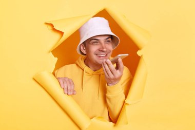 Sıradan kapüşonlu ve panama giyen mutlu bir adamın portresi sarı kağıttaki yırtık deliğe bakarak sanal asistan, dijital hoparlör uygulaması kullanarak cep telefonuyla konuşuyor..