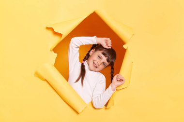 Portrait of funny positive little girl with braids wearing casual shirt looking through torn hole in yellow paper, keeps hands raised, having fun, expressing happiness.
