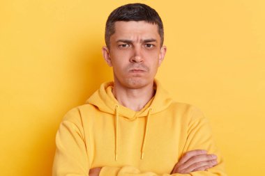 Üzgün sinirli genç yetişkin bir adam, günlük tarz kapüşonlu, sarı arka planda yalnız poz veriyor, katlanmış ellerle duruyor, kameraya bakıyor, kederini ifade ediyor..