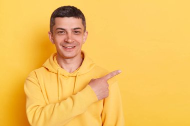 Gülümseyen iyimser genç bir adamın portresi günlük tarz kapüşonlu, sarı arka planda izole edilmiş poz veriyor, ayakta duruyor ve işaret ediyor, reklam alanını gösteriyor..