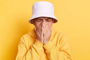 Üzücü adam, günlük tarz kapüşonlu ve panamalı, kederli, kötü ruh hali, sorunları, depresyonu olan, ağlayan, yüzünü palmiyelerle örten, sarı arka planda izole edilmiş poz veren üzgün bir adam..