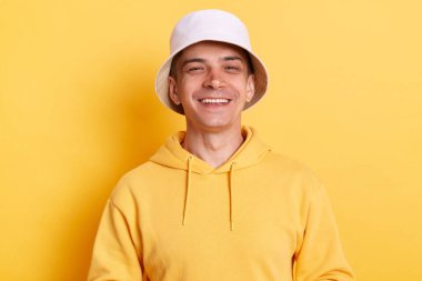 Keyifli beyaz bir adamın portresi günlük tarz kapüşonlu ve panama giyen, kameraya olumlu bir ifadeyle bakan, mutlu olan, sarı arka planda izole edilmiş poz veren.