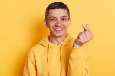 İyimser gülümseyen bir adamın portresi günlük tarz kapüşonlu, Kore 'nin kalp işaretini gösteren, sevgi, ilgi, barış veya desteğin sembolü, sarı arka planda izole edilmiş poz veren.