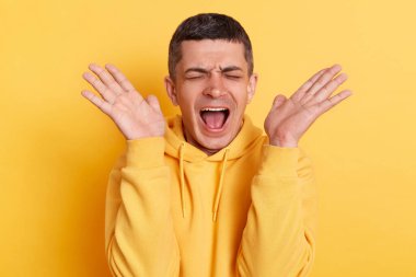 Kapalı mekanda, sarı arka planda izole edilmiş, mutsuz, kollarını kaldırmış bağırarak bağıran, sıradan tarzda kapüşonlu giyinmiş, umutsuz esmer bir adamın fotoğrafı..