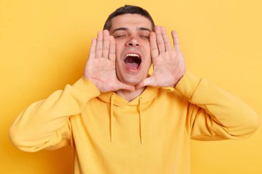 Sarı arka planda yalnız poz veren, elleri ağıza yakın duran ve çığlıklar atan, önemli bilgileri anons eden, günlük tarz kapüşonlu bir adam..