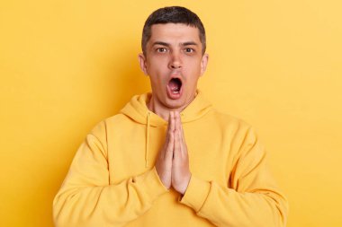 Şok olmuş, sıradan bir kapüşonlu giyen, sarı arka planda izole edilmiş yoga pozu veren ve rahatlamaya çalışan bir adamın portresi..