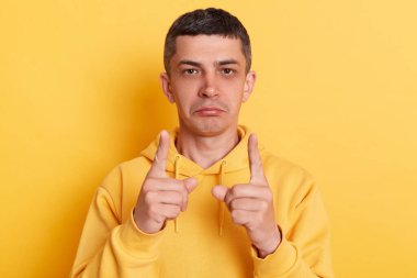 Sarı arka plan üzerinde izole edilmiş, küçük bir jest, küçük bir miktar ya da düşük ölçekli işaret gösteren, günlük tarz kapüşonlu bir adam kapalı çekim.