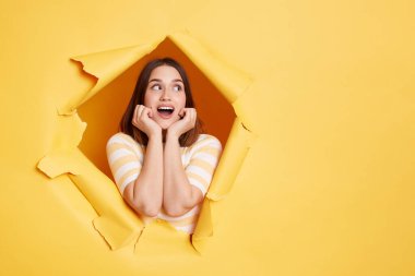 Şaşırmış esmer kadının kapalı mekanda çekilmiş fotoğrafı yırtık kağıt delikte duruyor, ağzı açık bakıyor, ellerini çenesinin altında tutuyor, sarı arka planda çığır açacak bir şey arıyor..