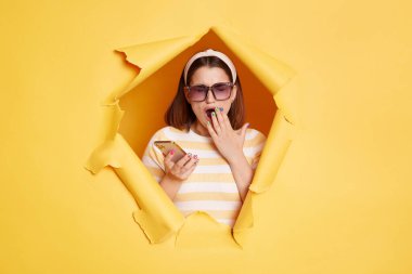 Yorgun esmer kadının portresi yırtık kağıt delikte duruyor, sarı arka planda çığır açıyor, cep telefonu tutuyor, esniyor, sıkılıyor ve telefon kullanmaktan yoruluyor..