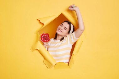 Mutlu, neşeli, çizgili gömlek ve saç bandı takan bir kadının kapalı çekimleri. Keyfi yerindeyken uyanıyor, gülümsüyor ve ellerini uzatıyor. Sarı kağıt arka planda beliriyor..