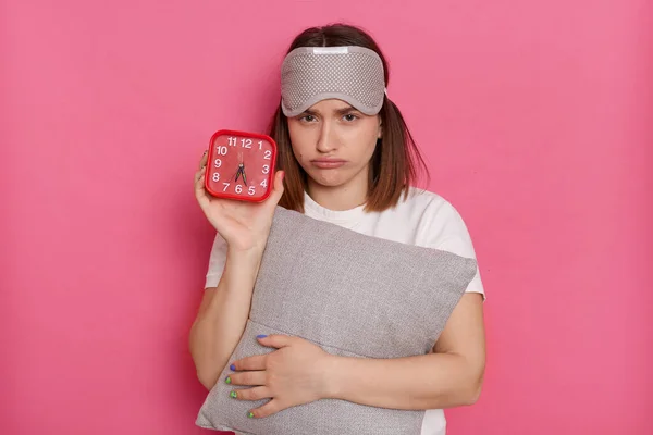 Caucasian young woman with red alarm clock and pillow awakening hard early, posing isolated on pink background, having no enough sleep, want more rest at weekends.