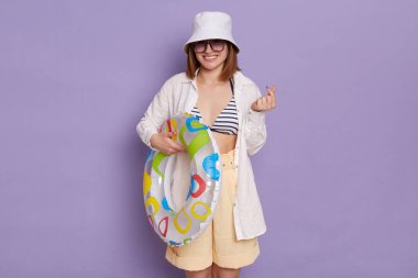 İyimser kadın Koreli kalp işareti, sevgi, ilgi, barış ya da destek sembolü, mayo, gömlek, şort ve panama giymiş, mor arka planda izole edilmiş poz veriyor..