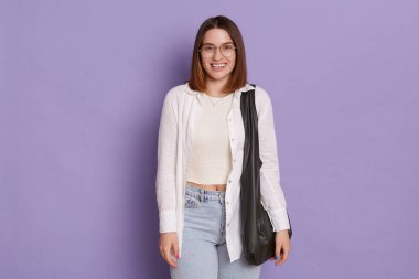 Yatay olarak gülümseyen, siyah çantalı, beyaz gömlekli, kot pantolonlu, mor arka planda izole edilmiş bir kadın, kameraya bakıyor, pozitif duyguları ifade ediyor, mutlu..