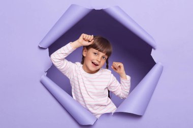 Kapalı alanda, örgülü, çizgili tişörtlü, yırtık kağıt duvarda poz veren, komik bir yüz ifadesi olan kameraya bakan, kollarını kaldırıp dans eden iyimser küçük bir kız resmi..