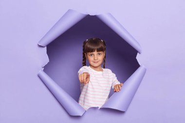 Çizgili gömlek giyen ciddi küçük bir kızın portresi mor kağıt arka planda kırılıyor, kadın çocuk parmak ve kamerayı gösteriyor, seni seçiyor, seçimini gösteriyor..