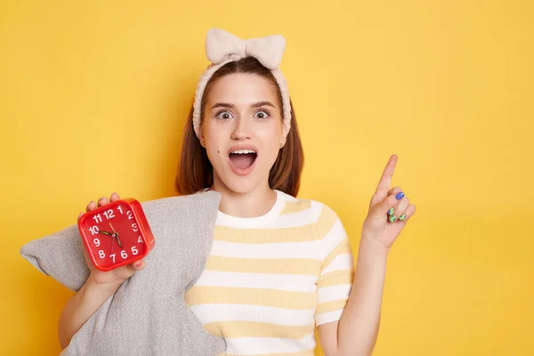 Portrait of excited woman wearing t shirt and hair band posing isolated on yellow background, holding pillow and red alarm clock, looking at camera with excitement, pointing aside at copy space.