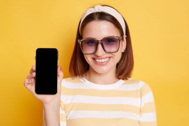 Sarı arka planda izole edilmiş çizgili tişört giyen neşeli, koyu renk saçlı genç bir kadının kapalı çekimleri, reklam için boş ekranlı cep telefonunu gösteriyor..