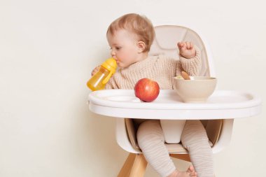 Bej renkli kazak giyen, sarı şişeden su içen, etrafını kolaçan eden, beyaz arka planda izole edilmiş poz veren susamış bir bebek resmi.