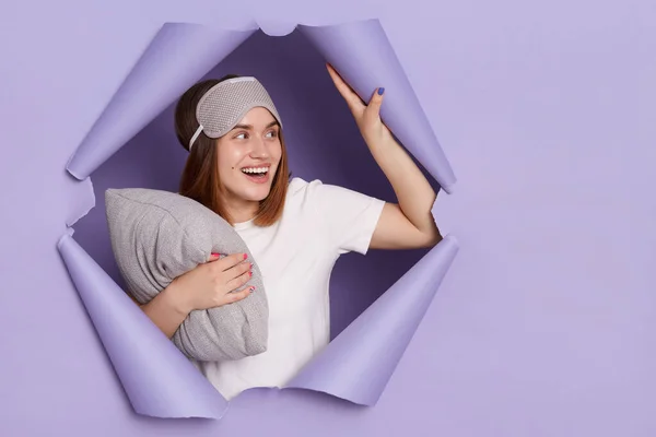 Indoor shot of excited dark haired woman wearing t shirt and blindfold standing in purple paper hole with pillow in hands, looking away at copy space for promotional text.