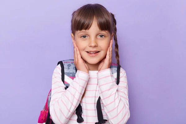 Portrait of positive smiling Caucasian little girl with braids touching her cheeks, wearing backpack and looking at camera with positive expression, posing isolated on purple background.