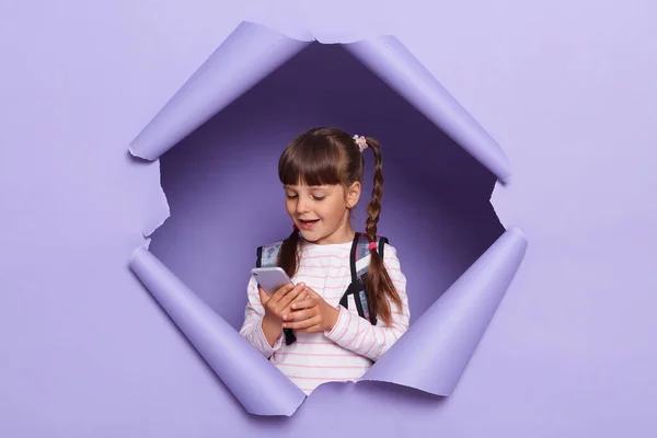 Wow, cool mobile app. Portrait of excited Caucasian school girl wearing backpack, holding and using smartphone, looking at gadget screen, posing in purple paper hole.
