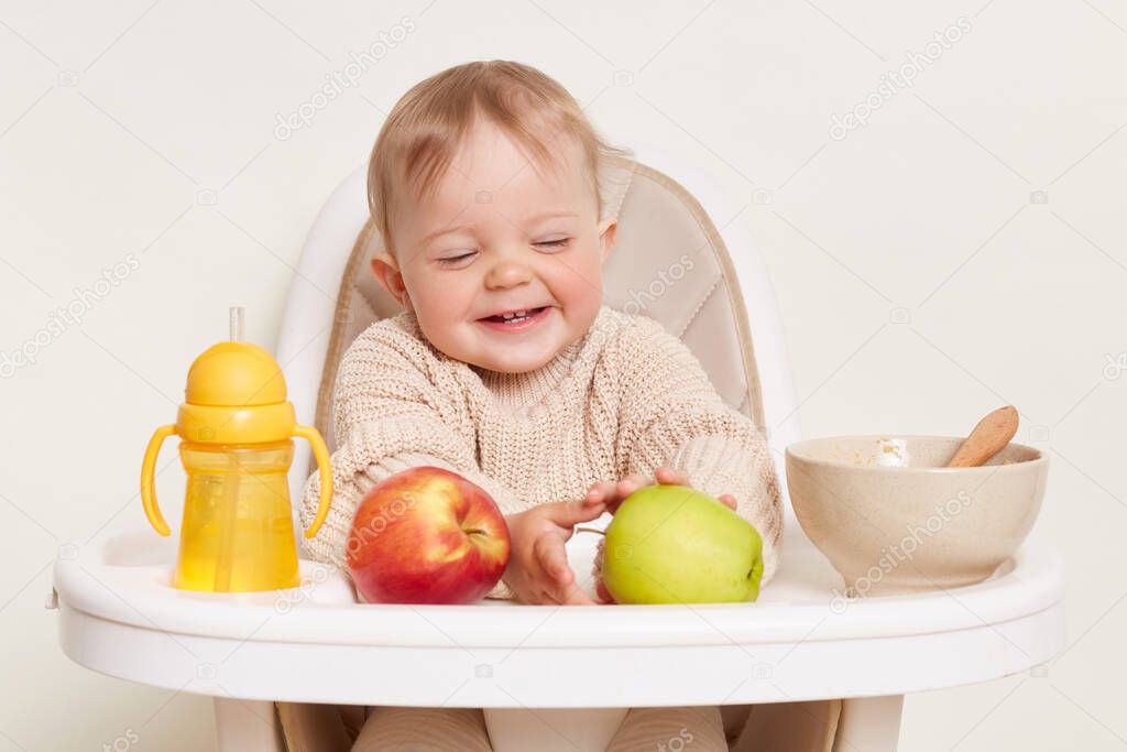 happy smiling cute little female baby kid wearing beige sweater sitting in high chair, finishing her porridge and wants to eat apple, isolated over white background.