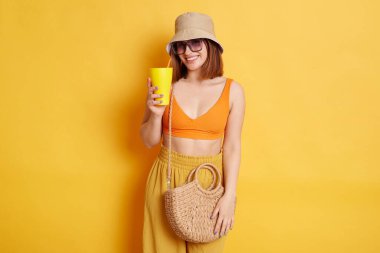 Güneş gözlüğü takan, sarı arka planda izole edilmiş poz veren, elinde lezzetli taze içeceklerle kameraya gülümseyen neşeli, koyu renk saçlı bir kadının portresi..
