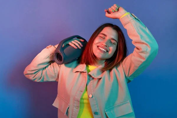 Horizontal shot of satisfied delighted woman wearing stylish jacket posing isolated on neon light background, holding musical column and and dancing, enjoying music.