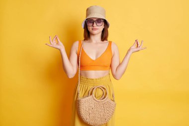 Rahatlamış beyaz kadın portresi mayo, şort ve panama giyen, sarı arka planda izole edilmiş poz veren, yoga yapan, rahatlatıcı.