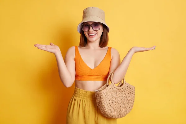 Horizontal shot of positive young young woman wearing swimsuit, shorts and panama, posing isolated over yellow background, standing spread arms, being not sure, don\'t know.