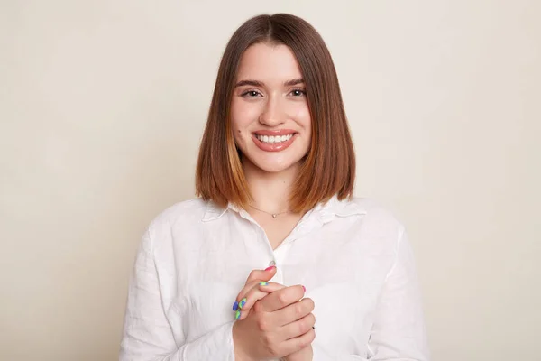 Indoor shot of positive optimistic dark haired woman wearing shirt posing isolated over white background, looking at camera with positive expression, keeps hands together.