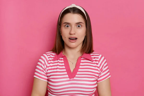 Horizontal shot of shocked surprised woman wearing striped T-shirt and hair band posing isolated over pink background, looking at camera with big eyes and open mouth.