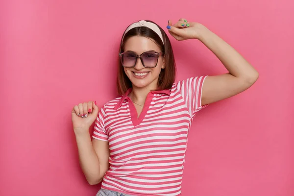 Horizontal shot of attractive playful happy woman wearing striped T-shirt and sunglasses, standing with raised arms, dancing, celebrating holiday, posing isolated over pink background.