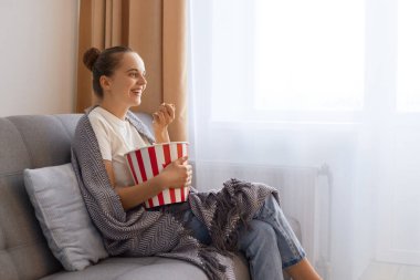 Kanepede patlamış mısırla oturan, film izleyen, patlamış mısır yiyen, boş zamanın tadını çıkaran, gülümseyen ve komediden zevk alan genç bir kadının yan görüş portresi..