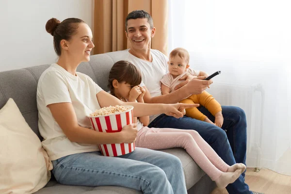 Side view of happy family watching television while eating popcorn together on the sofa, mother holding remote choosing movie or cartoon to watch, posing near window in living room.