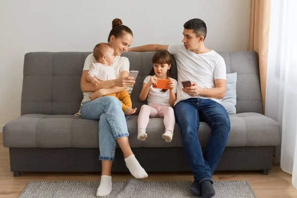 Portrait of family mother father and two daughters holding and using their mobile phones, looking at daughter\'s phone, spending time checking social networks or reading news, or watching cartoons.